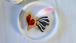 Decorative brooch with red heart and black and white striped bow on a white plate. The feather is painted with a paint gradient. 
